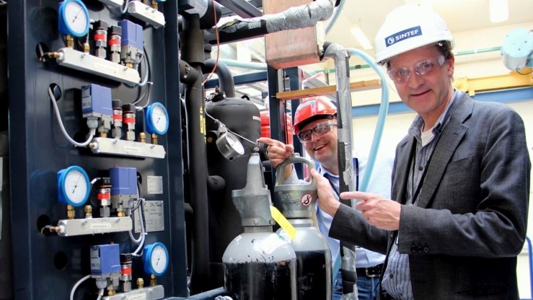 Two people wearing safety goggles and helmets, one with a SINTEF logo, are standing by a technical installation with measuring instruments and valves.